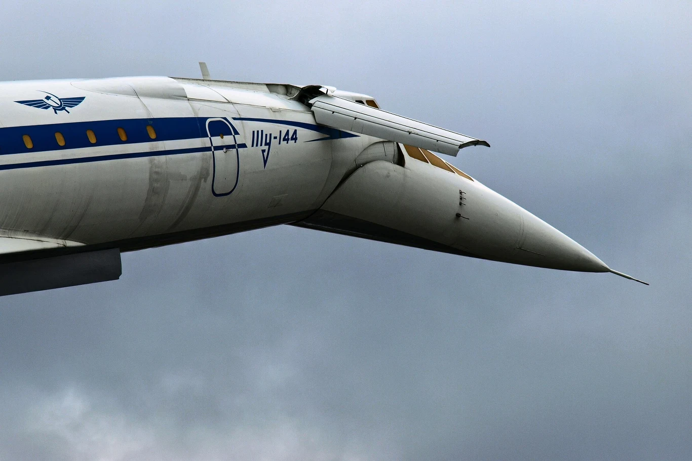Przód samolotu naddźwiękowego Tu-144 z wydłużonym nosem i opuszczonym skrzydełkiem ogonowym na tle chmur.