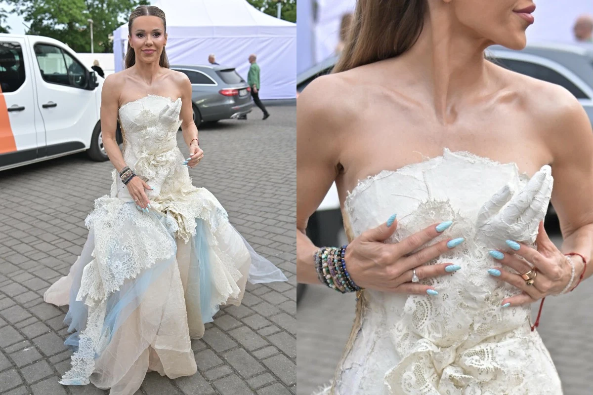 Doda w modnym manicure na festiwalu w Opolu Kobieta w białej, koronkowej sukni o nieregularnym kroju z delikatnymi niebieskimi akcentami, z długimi, jasnymi włosami oraz niebieskimi paznokciami, poprawia kreację dłońmi ozdobionymi bransoletkami, stojąc na brukowanej nawierzchni.