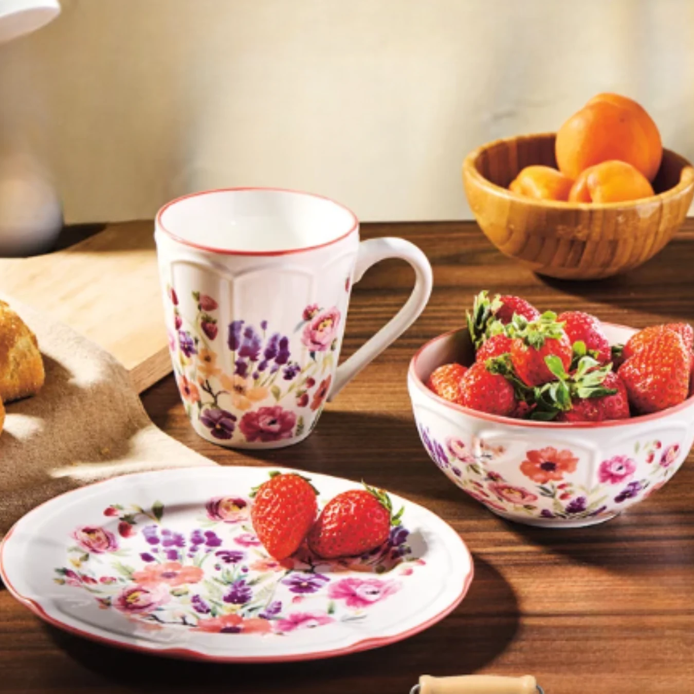 Ceramiczny zestaw naczyń z kwiatowym wzorem, składający się z talerzyka, kubka i miseczki wypełnionej truskawkami, obok koszyczek z morelami.