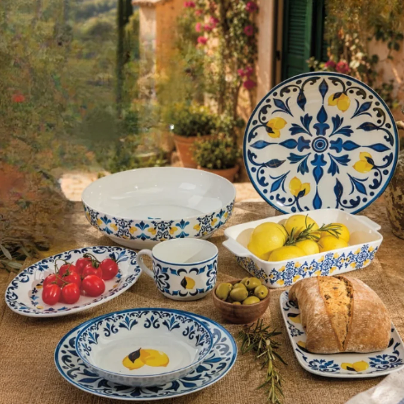 Zestaw ceramicznych naczyń z kolorowym, kwiatowym wzorem, ułożonych na lnianym obrusie, z dodatkiem świeżych pomidorów, cytryn, oliwek i pieczywa.