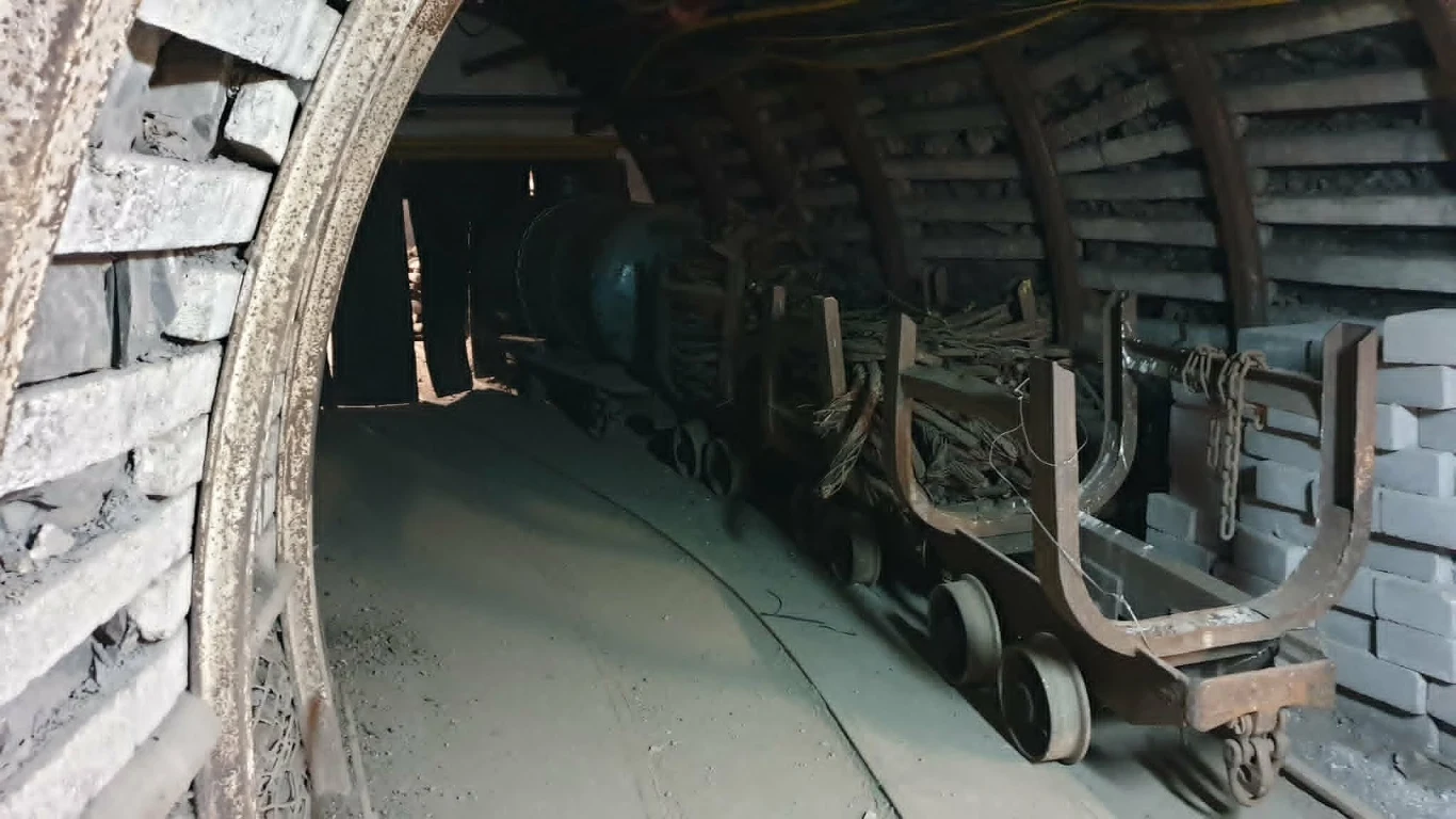 Wąski tunel z metalową konstrukcją wsporczą i torami, na których stoją puste, zabrudzone wózki kopalniane oraz zwinięte przewody obok stosu betonowych bloków.