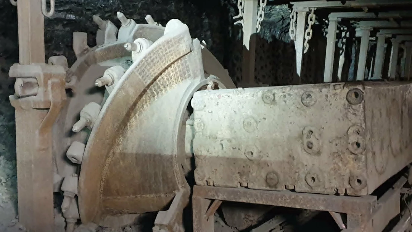 Duża maszyna górnicza z masywną tarczą tnącą w otoczeniu metalowych elementów konstrukcyjnych i ciemnych skał.