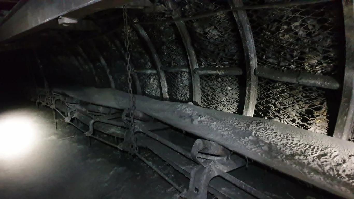 Przemysłowy przenośnik taśmowy pokryty pyłem w wąskim, półkolistym tunelu zabezpieczonym metalowymi wspornikami i siatką ochronną.
