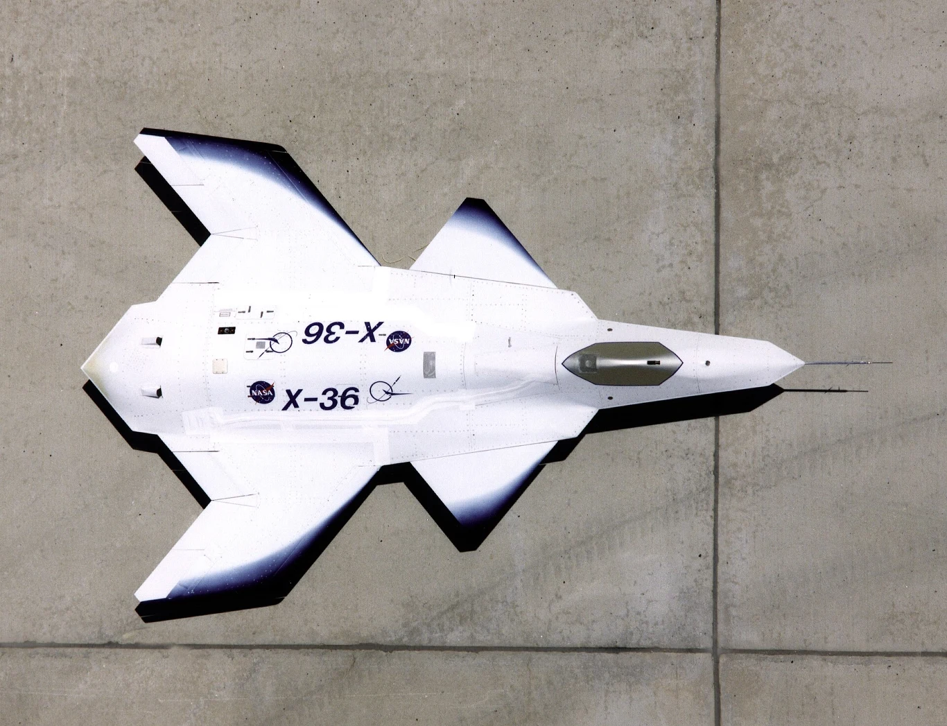 X-36 Eksperymentalny bezzałogowy samolot typu X-36 o futurystycznym kształcie, z charakterystycznymi powłokami w biało-czarnej kolorystyce oraz logo NASA na kadłubie, sfotografowany z góry na betonowym podłożu.