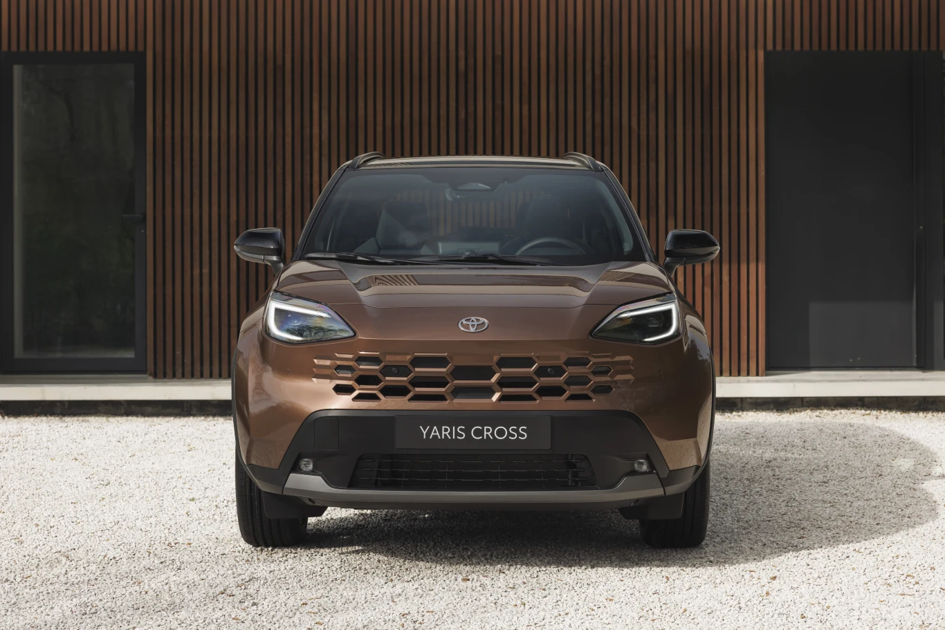 Toyota Yaris Cross najbardziej zmieniła się z przodu Brązowa Toyota Yaris Cross po liftingu, widok z przodu, grill i reflektory LED, tło: drewniane panele fasady.