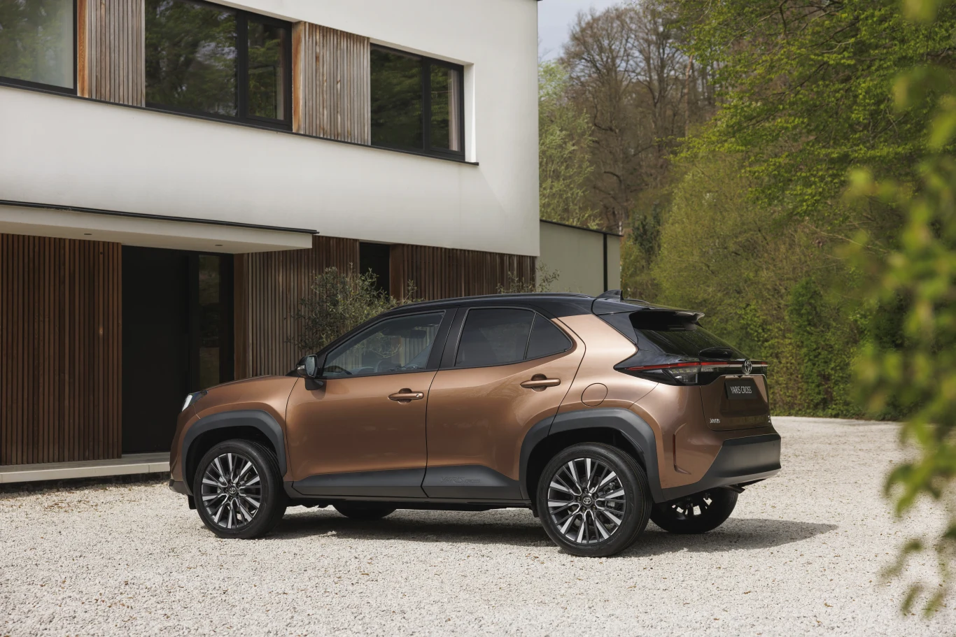 Toyota Yaris Cross Brązowy SUV zaparkowany na żwirowym podjeździe obok nowoczesnego budynku z dużymi oknami i drewnianymi detalami, w otoczeniu zieleni.