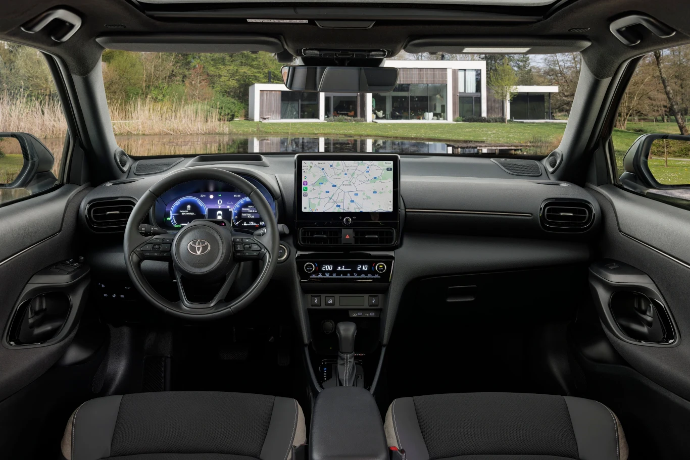 Toyota Yaris Cross Nowoczesne wnętrze samochodu marki Toyota z cyfrowym wyświetlaczem za kierownicą oraz dużym ekranem dotykowym na środkowej konsoli, przez przednią szybę widoczny nowoczesny dom otoczony zielenią.