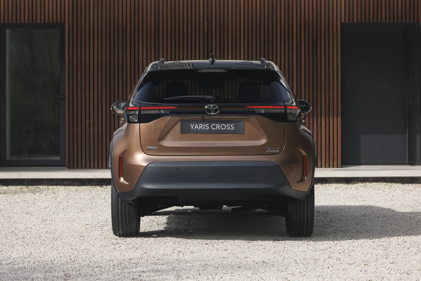 Toyota Yaris Cross Brązowa Toyota Yaris Cross widoczna od tyłu, zaparkowana na jasnym żwirze na tle elewacji z poziomych drewnianych paneli i dwóch okien.