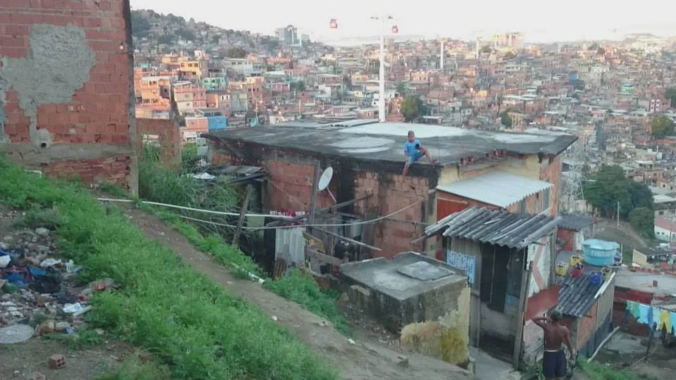 Widok na Complexo do Alemao, jedną z największych faweli w Rio de Janeiro Zabudowa z czerwonej cegły z prowizorycznymi dachami, dzieci bawiące się na dachu, rozległe, gęsto zaludnione osiedle w tle.