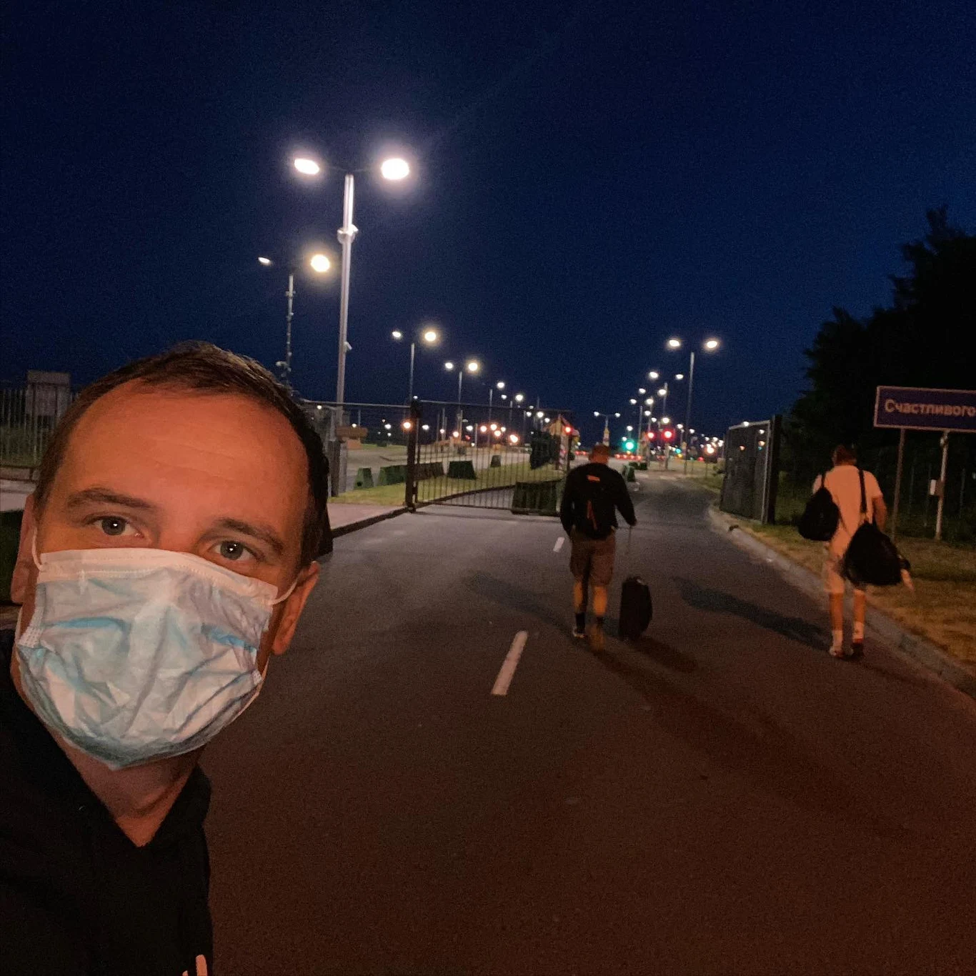 Przekraczanie granicy rosyjsko-polskiej jako "mrówka". Maseczka w tamtych czasach była obowiązkowa Mężczyzna w maseczce spogląda w kierunku aparatu, a dwóch innych ludzi z walizkami idzie oświetloną ulicą po zmroku.