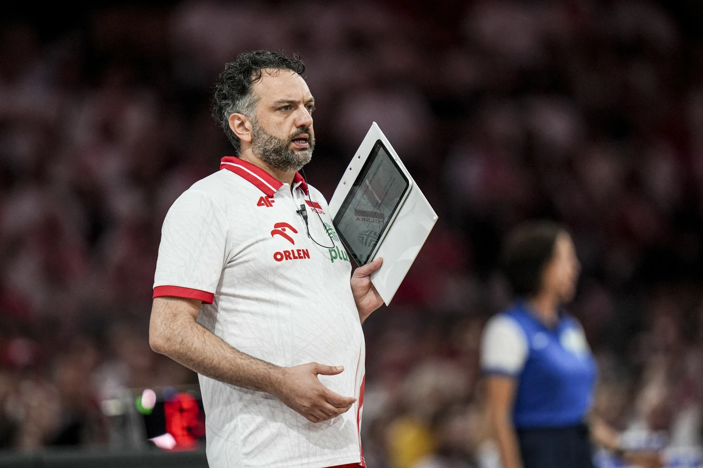Trener ubrany w biało-czerwoną koszulkę z logo sponsora i napisem Polska trzymający tablicę taktyczną podczas meczu siatkówki.
