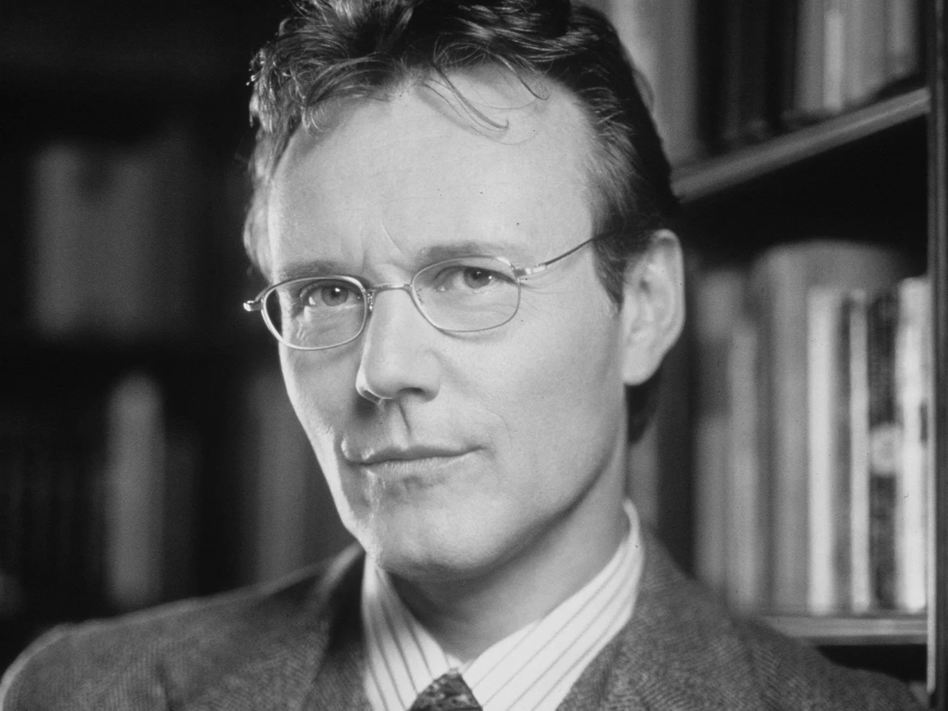 Anthony Head jako Rupert Giles w serialu "Buffy: Postrach wampirów" Mężczyzna w okularach i marynarce, stojący na tle półki z książkami.