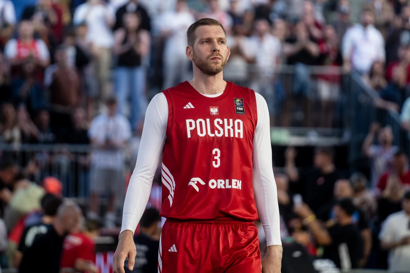Koszykarz w czerwonym stroju reprezentacji Polski z numerem 3, stojący na boisku w otoczeniu widowni.