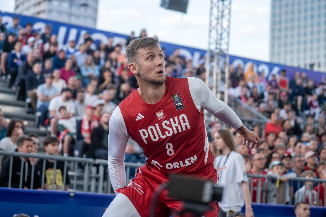 Koszykarz w czerwonym stroju z napisem 'Polska' i numerem 8 podczas meczu na otwartym boisku, otoczony przez widownię na trybunach.