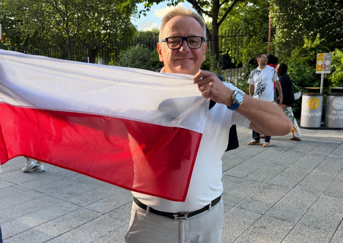 Mężczyzna z okulami i jasnymi włosami trzyma rozwiniętą polską flagę na tle zielonych drzew i przechodniów.