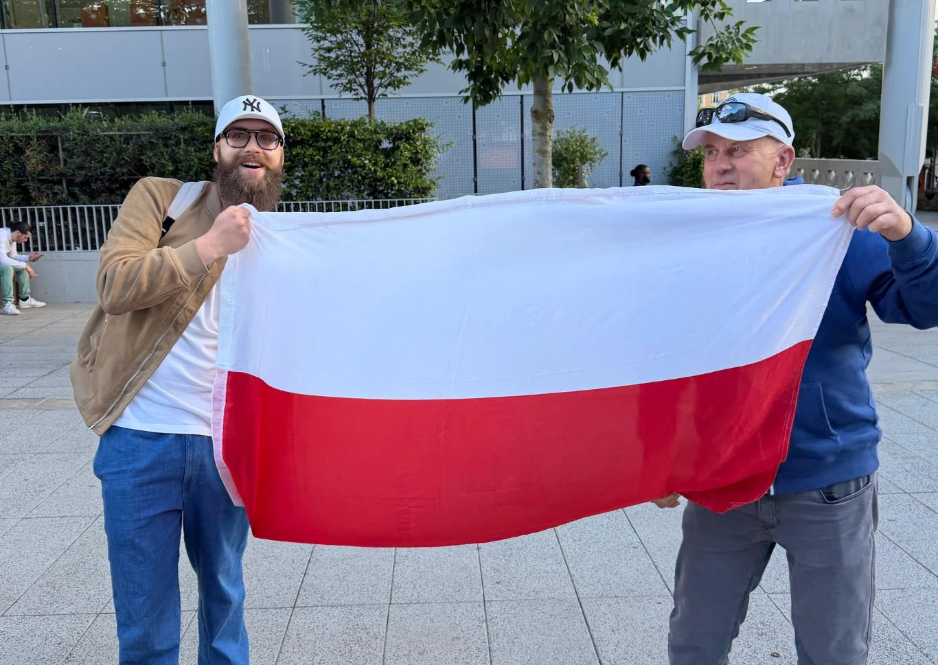 Dwaj mężczyźni trzymają rozwiniętą biało-czerwoną flagę Polski, stojąc obok siebie na chodniku w miejskim otoczeniu.