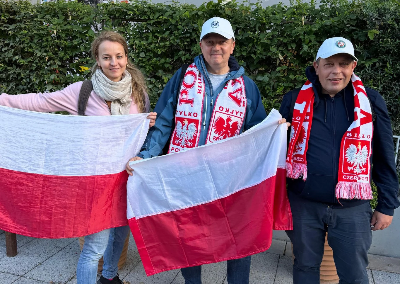 Trzy osoby w biało-czerwonych barwach trzymają flagi Polski i mają na sobie szaliki kibica z polskimi symbolami.