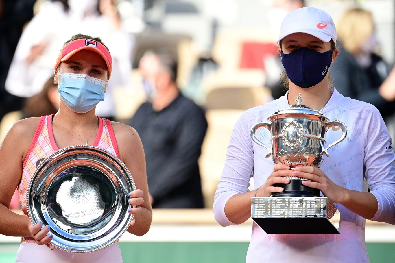 Dwie tenisistki w sportowych strojach i maseczkach ochronnych stoją obok siebie, trzymając nagrody – jedna srebrną paterę, druga okazały puchar turniejowy.