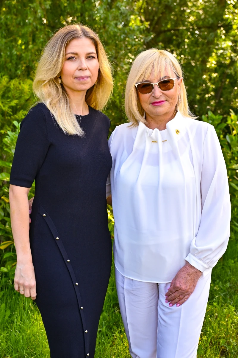 Dwie kobiety stoją obok siebie w zielonym otoczeniu; jedna ubrana w elegancką czarną sukienkę, druga w białej bluzce i spodniach, z okularami przeciwsłonecznymi.