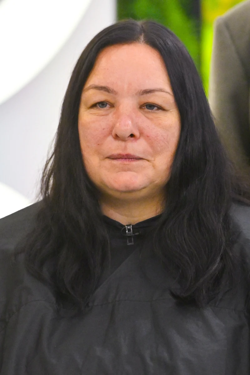 Justyna Górka przed metamorfozą Kobieta o długich, czarnych włosach i naturalnym wyglądzie twarzy, ubrana w ciemne pelerynę fryzjerską, stoi na tle jasnych i zielonych elementów.