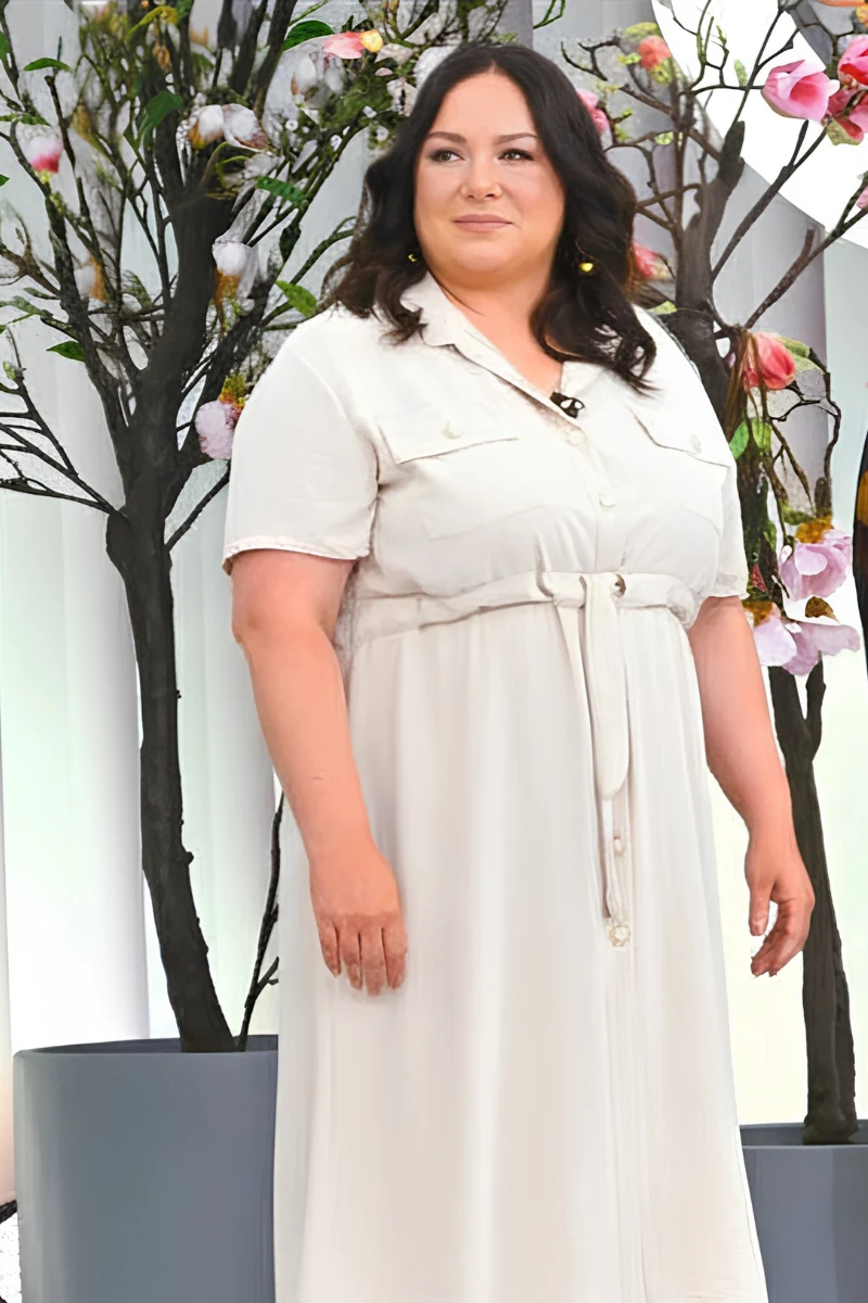 Justyna Górka po metamorfozie Kobieta w jasnej, długiej sukience z krótkim rękawem stoi na tle ozdobnych drzew z kwitnącymi gałązkami.
