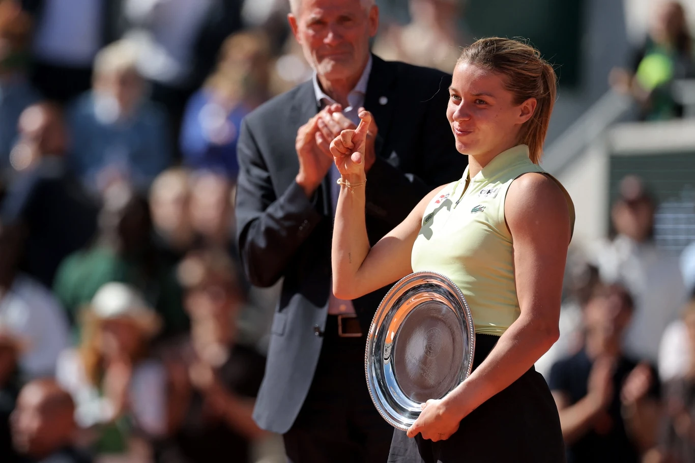 Maja Chwalińska po finale Rolanda Garrosa Kobieta trzymająca srebrny puchar w dłoni, uśmiechnięta, w sportowym stroju, z gratulującym jej mężczyzną w tle.