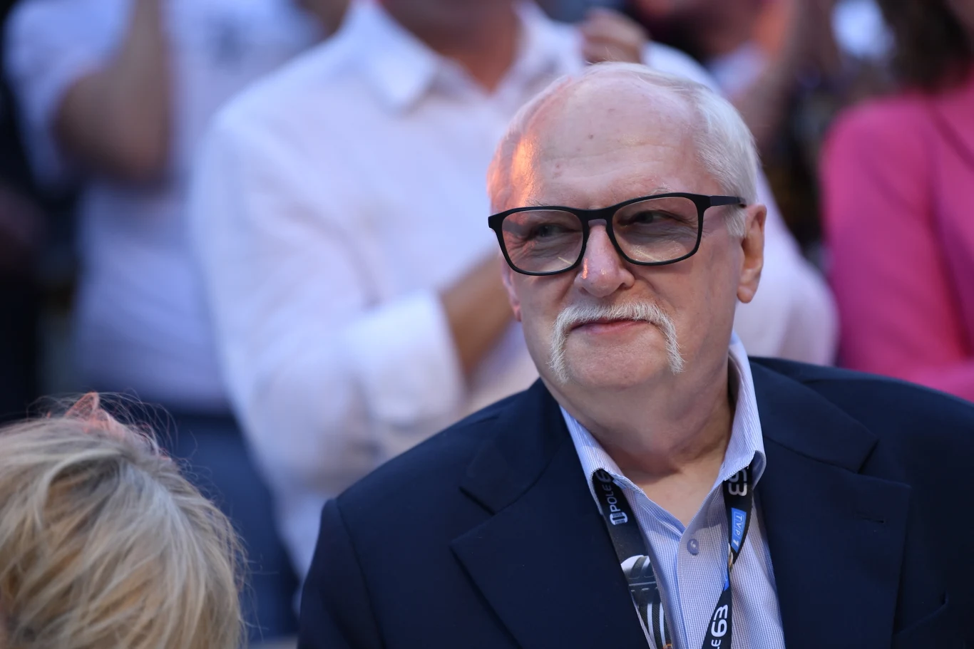 Starszy mężczyzna z białymi włosami i charakterystycznym wąsem, w okularach oraz eleganckiej marynarce, stoi w tłumie ludzi ubranych półformalnie.