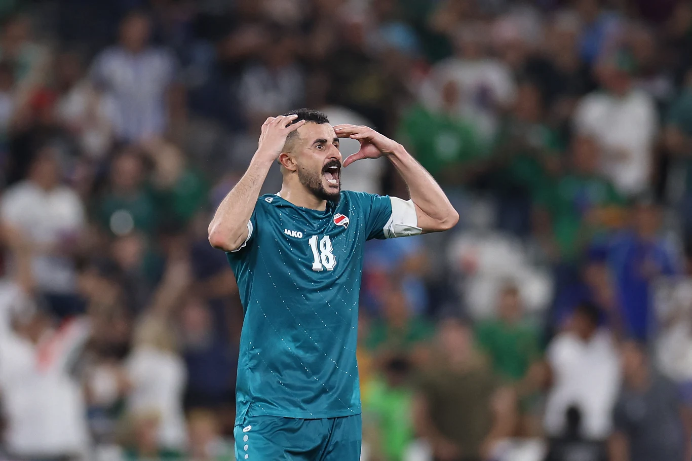Piłkarz w turkusowym stroju z numerem 18 i opaską kapitana wyraża silne emocje, trzymając ręce na głowie podczas meczu na stadionie wypełnionym kibicami.