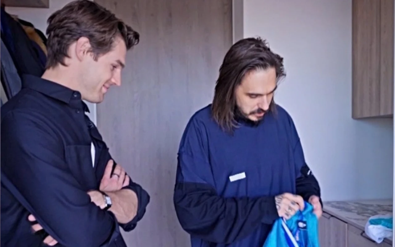 Dwóch mężczyzn w pomieszczeniu, jeden z nich trzyma w rękach niebieską koszulkę sportową i uważnie się jej przygląda, podczas gdy drugi obserwuje go z uśmiechem.