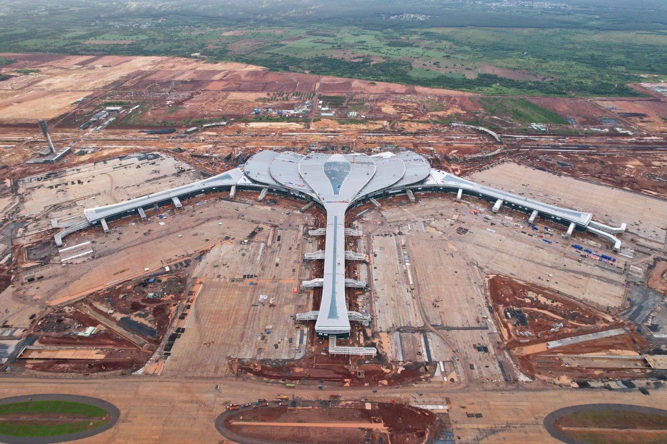 Nowoczesny terminal lotniska o charakterystycznym, rozgałęzionym kształcie otoczony rozległym placem budowy, z trzema głównymi ramionami odchodzącymi od centralnej części budynku.