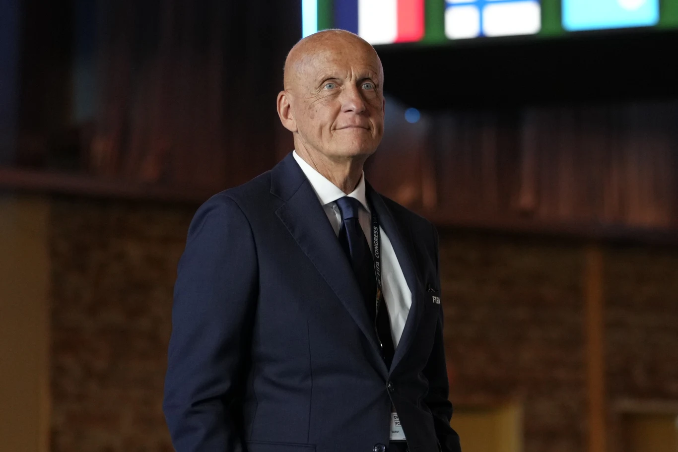 Łysy mężczyzna w eleganckim, granatowym garniturze i krawacie stoi w dobrze oświetlonej sali z widocznym fragmentem kolorowych flag na ekranie w tle.
