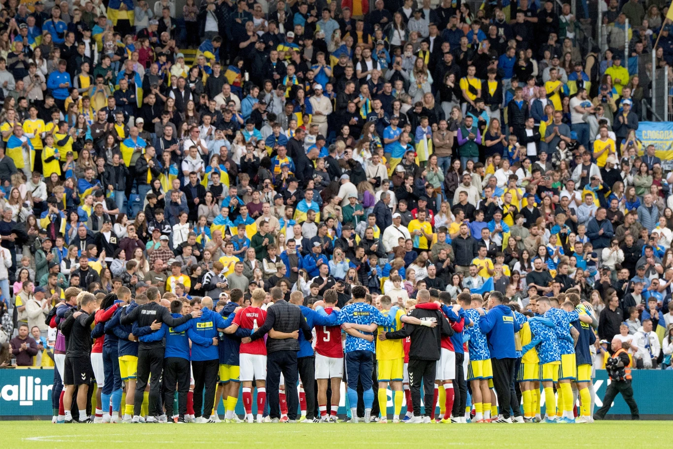 Zawodnicy i sztab szkoleniowy zebrani w kręgu na murawie stadionu, otoczeni tłumem kibiców w żółto-niebieskich barwach na trybunach.