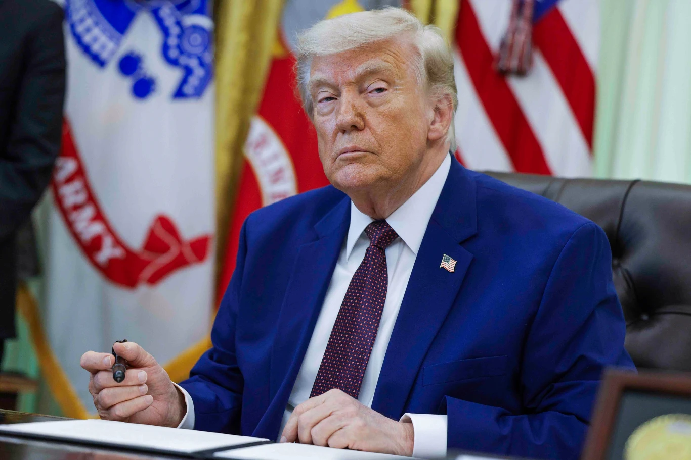 Donald Trump Donald Trump w granatowym garniturze siedzi przy biurku z dokumentami i długopisem, w tle flagi i zasłony.