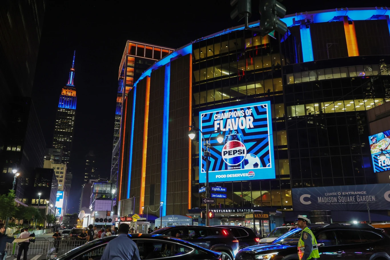 Madison Square Garden Kolorowo podświetlony budynek Madison Square Garden z dużym ekranem reklamującym Pepsi, otoczony samochodami i pieszymi na ruchliwej ulicy nocą; w tle wieża Empire State Building iluminowana różnobarwnym światłem.