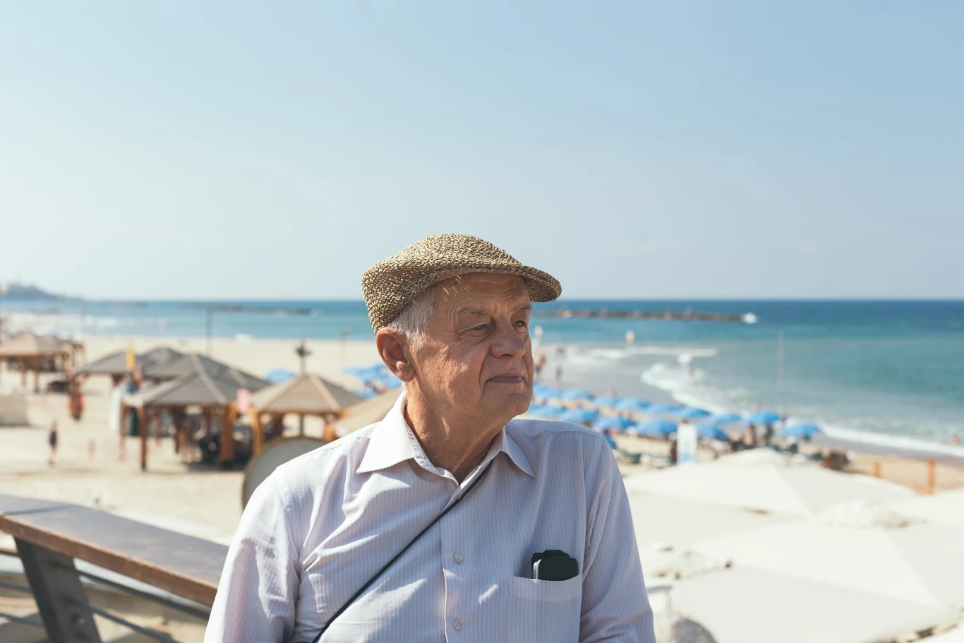 Starszy mężczyzna w jasnej koszuli i kaszkiecie stoi nad brzegiem morza, z tyłu widoczne są leżaki oraz parasole plażowe, a także spokojna, błękitna woda.