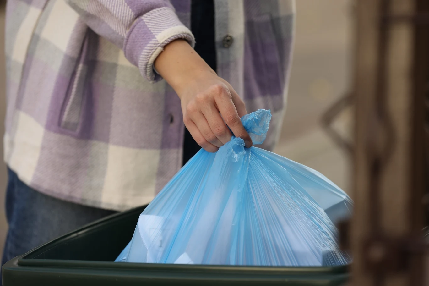 Ręka w kolorowej koszuli wrzuca niebieski worek na śmieci do zielonego kosza.