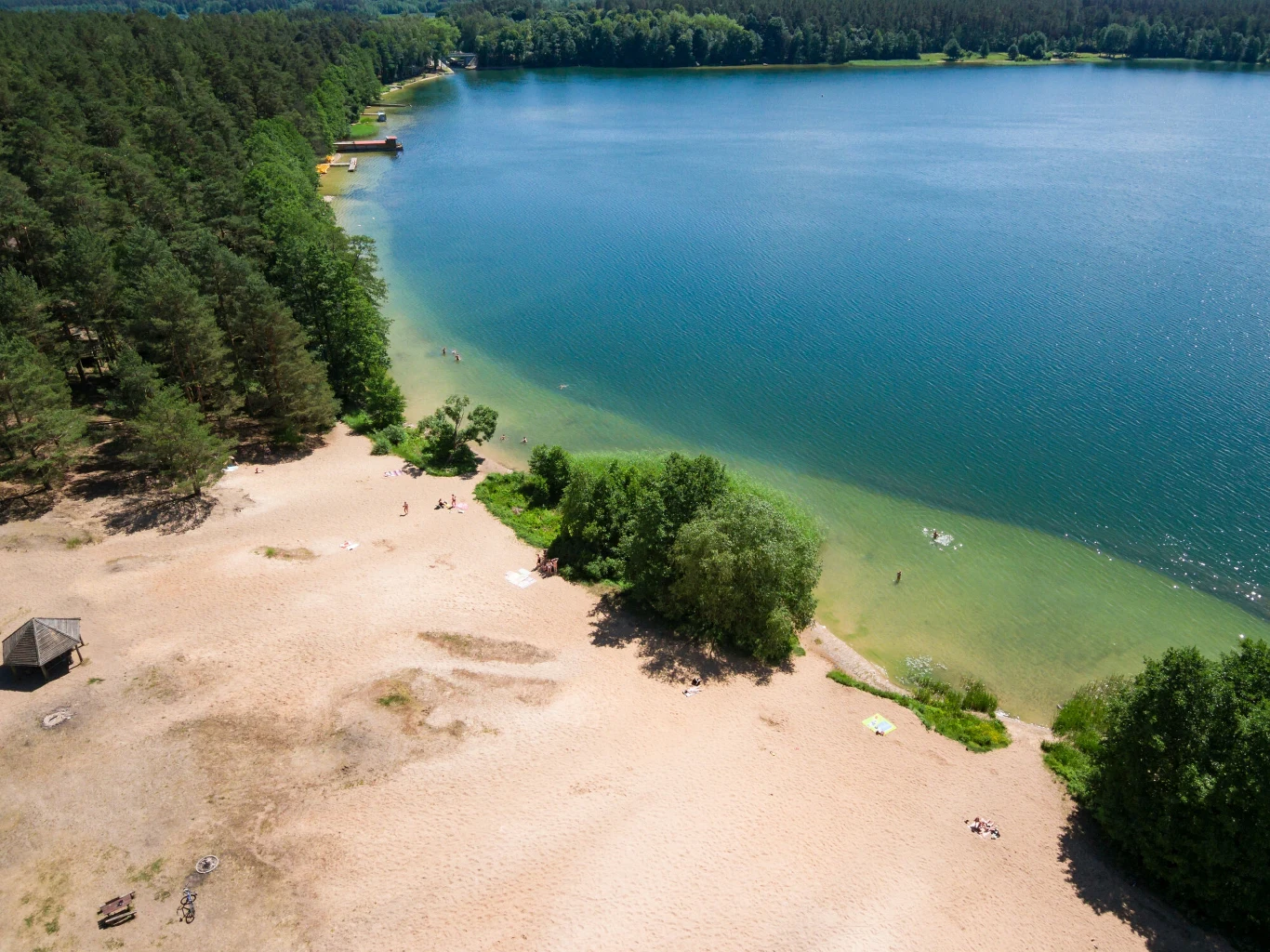 Piaszczysta plaża otoczona lasem, przechodząca w czystą, błękitną wodę jeziora; na brzegu rozłożone koce i kilka osób korzystających ze słonecznej pogody.