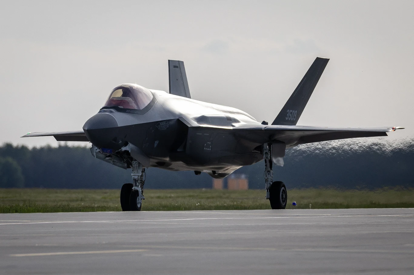 Myśliwiec F-35 ustawiony na pasie startowym z opuszczonym podwoziem, otaczający go pas zieleni i niebo zasnute chmurami.