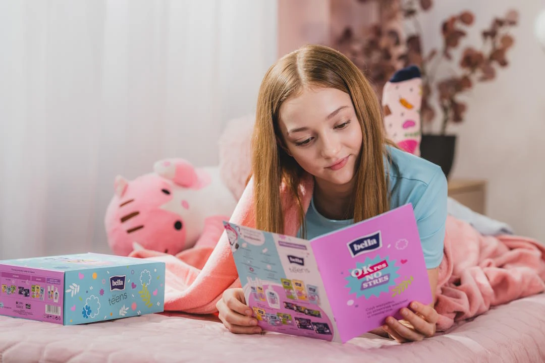 Dziewczyna leżąca na różowym łóżku przegląda kolorową ulotkę lub katalog marki Bella, obok leży pudełko tej samej marki oraz pluszowa zabawka w kształcie głowy Hello Kitty.