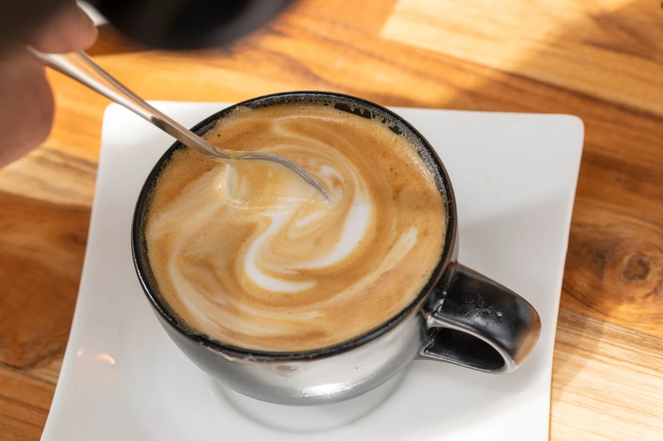 Wsyp łyżkę tego dodatku do kawy. Wzmocnisz stawy i skórę Czarna filiżanka cappuccino z mleczną pianką mieszana srebrną łyżeczką na białym talerzyku na drewnianym stole.