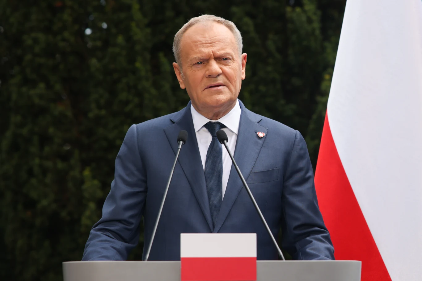 Premier Donald Tusk przemawia przy mównicy z polską flagą, obok duży biało-czerwony sztandar.