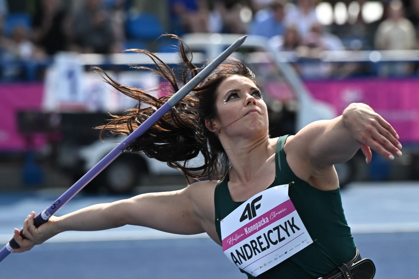 Zawodniczka w zielonym stroju sportowym wykonuje rzut oszczepem podczas zawodów lekkoatletycznych, skupiając wzrok przed siebie z dynamicznym ruchem ręki i ciała.