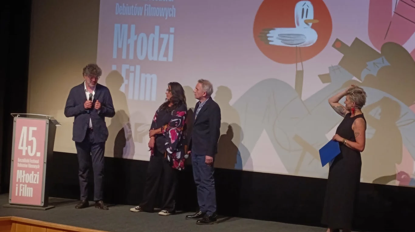 Czworo osób na scenie podczas wydarzenia filmowego, jedna z nich przemawia do mikrofonu, pozostali stoją obok, a w tle wyświetlony jest ekran z grafiką i napisem nawiązującym do festiwalu Młodzi i Film.