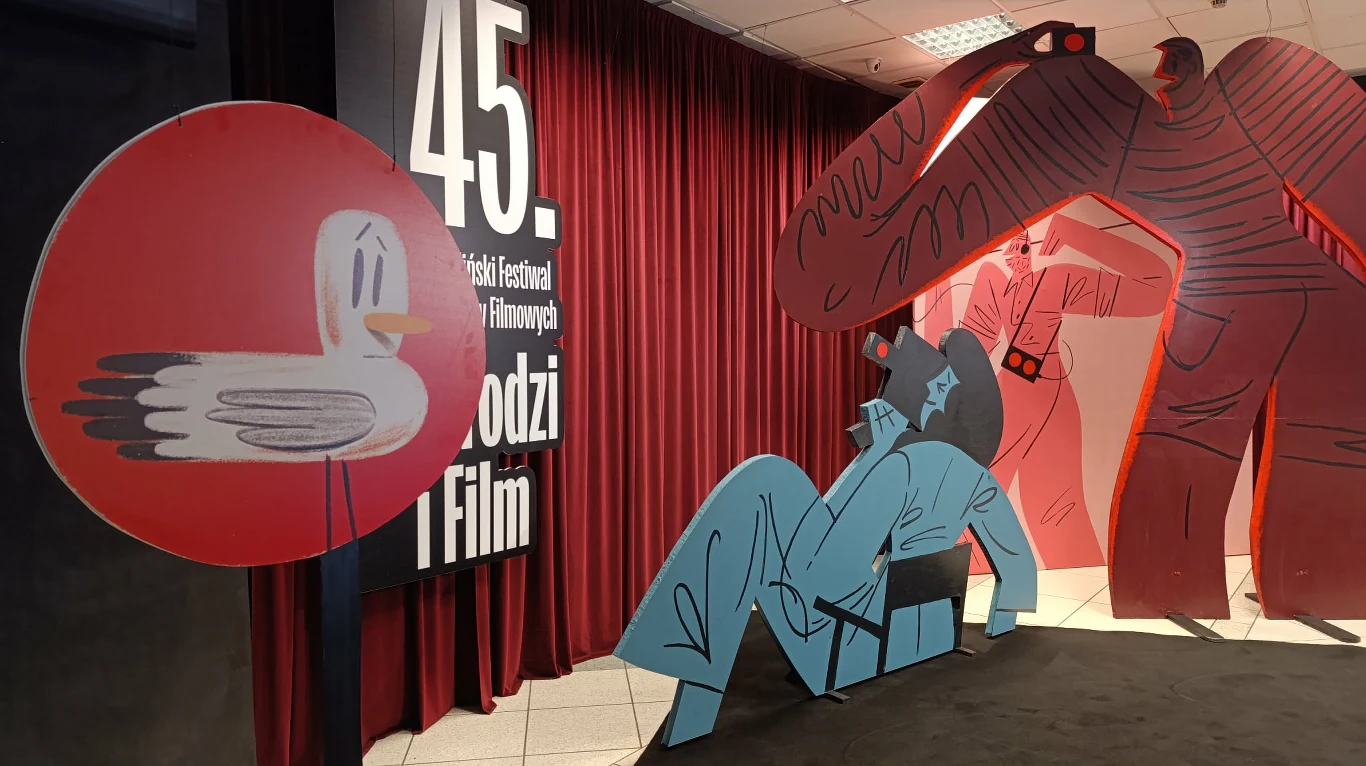 Duży baner z napisem „45. Festiwal Polskich Filmów Fabularnych Młodzi i Film” na tle czerwonej kurtyny, obok kolorowe, kartonowe postacie: niebieska postać leżąca, czerwona postać o dużych ramionach i okrągła tablica z rysunkiem mewy.