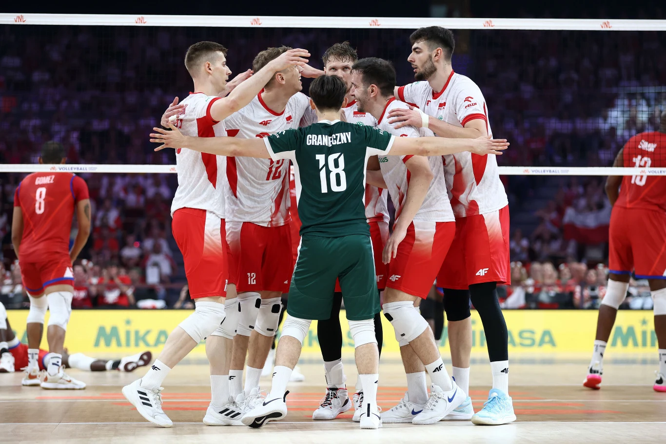 Grupa siatkarzy w biało-czerwonych strojach świętuje zdobycie punktu, stojąc razem w kręgu na boisku podczas meczu siatkówki, na pierwszym planie zawodnik w zielonym stroju z numerem 18.