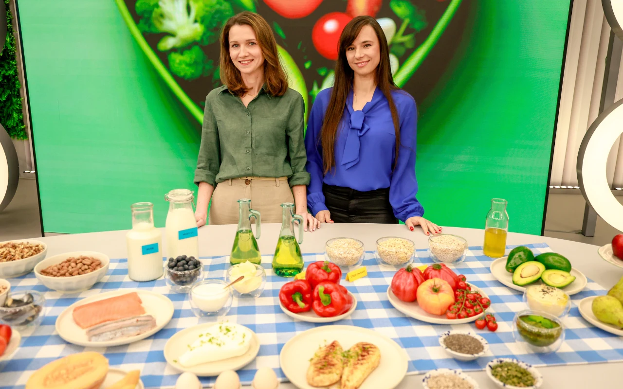 Dwie kobiety stoją przy stole zastawionym różnorodnymi produktami spożywczymi, takimi jak świeże owoce, warzywa, produkty mleczne oraz zdrowe przekąski, na tle dużego ekranu z zielonym motywem sałatki.