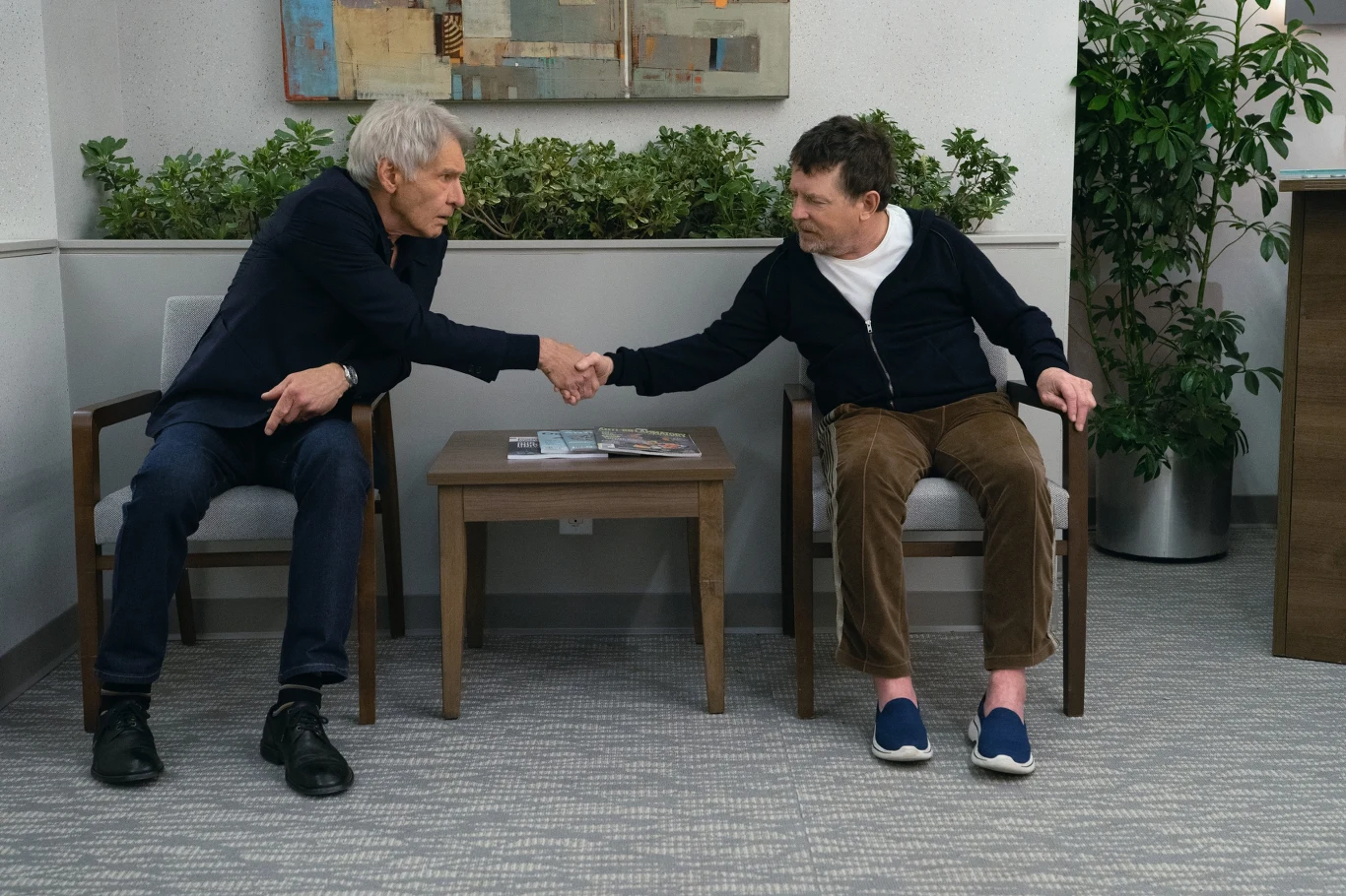 Harrison Ford i Michael J. Fox w serialu "Terapia bez trzymanki" Dwóch mężczyzn siedzi naprzeciwko siebie na krzesłach w nowoczesnym biurze lub poczekalni, ściskając sobie dłonie nad stolikiem z gazetami.