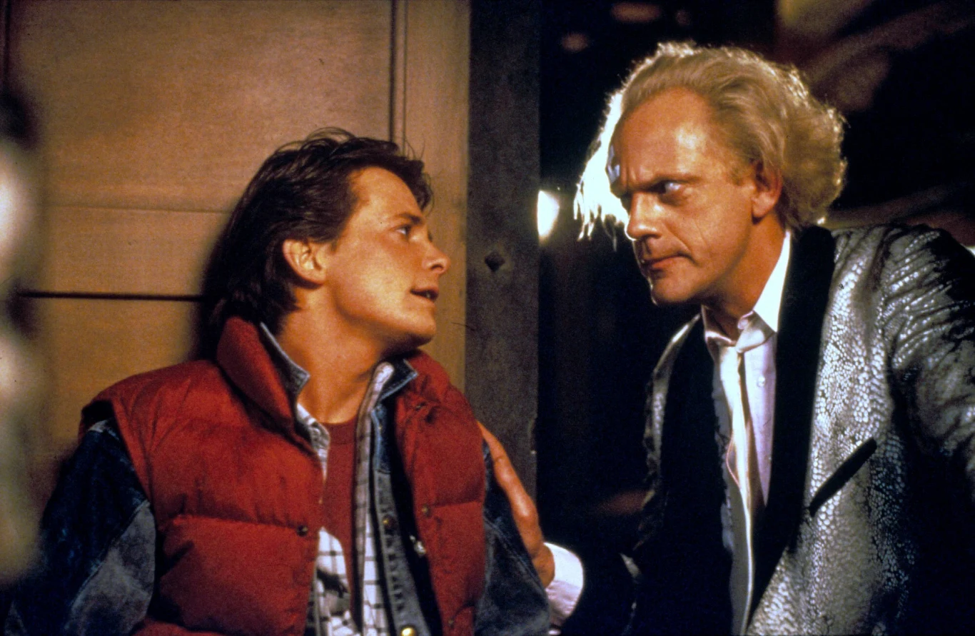 Michael J. Fox i Christopher Lloyd w filmie "Powrót do przyszłości" Dwóch mężczyzn w intensywnej rozmowie; jeden w czerwonej kamizelce i dżinsowej koszuli, drugi o siwych włosach w połyskującej marynarce, patrzący na siebie z powagą.
