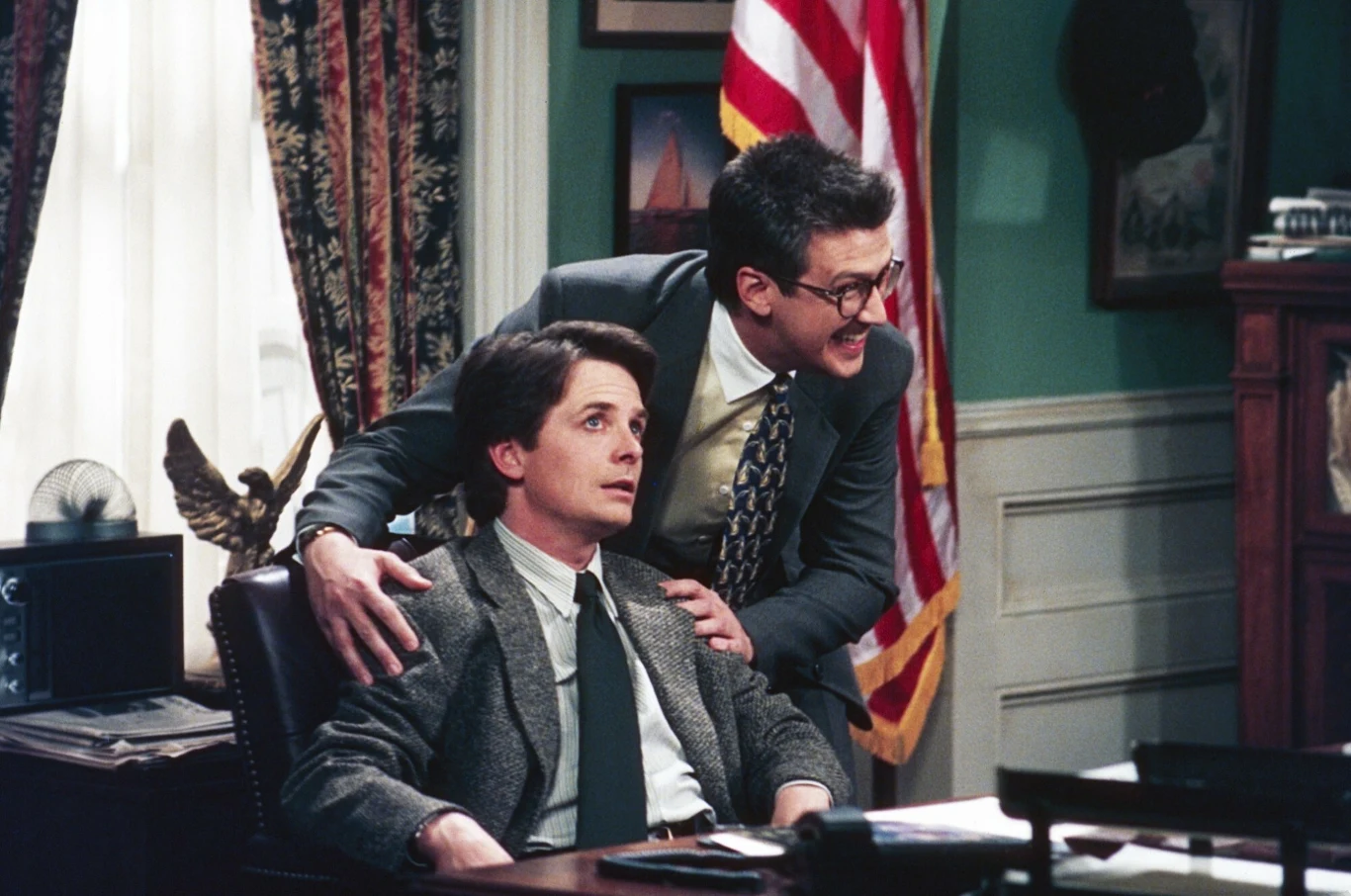 Michael J. Fox i Alan Ruck w serialu "Spin City" Dwóch mężczyzn w garniturach w gabinecie urzędowym; jeden siedzi za biurkiem z poważnym wyrazem twarzy, drugi stoi obok niego, opierając dłonie na jego ramionach, w tle amerykańska flaga.