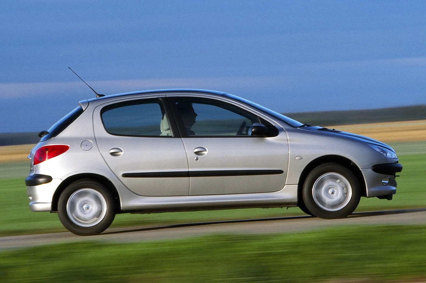 Srebrny hatchback poruszający się po wiejskiej drodze, z widocznym kierowcą za kierownicą.
